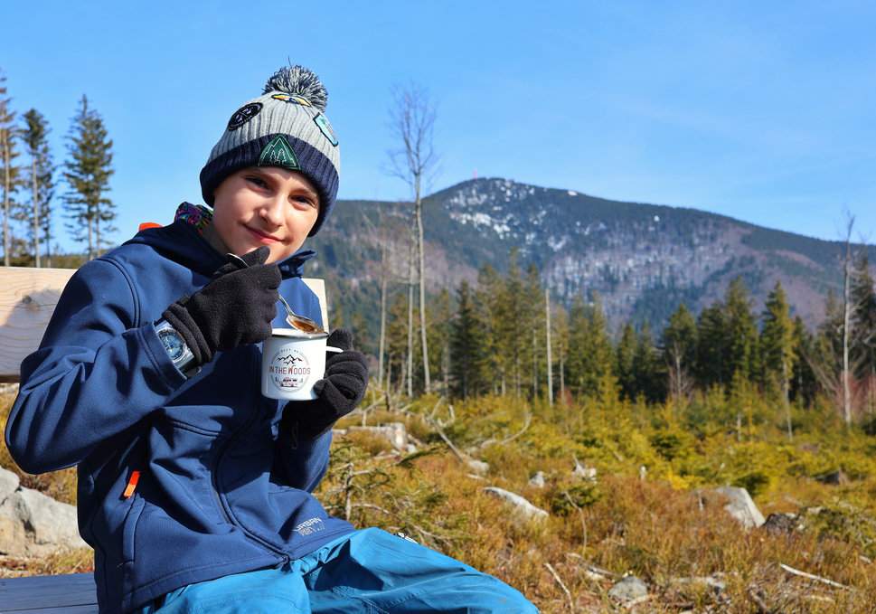 Szczęśliwe dziecko siedzące na drewnianej ławce, jedzące zupę z termokubka, w tle najwyższy szczyt Beskidu Morawsko-Śląskiego - punkt widokowy Smrčina