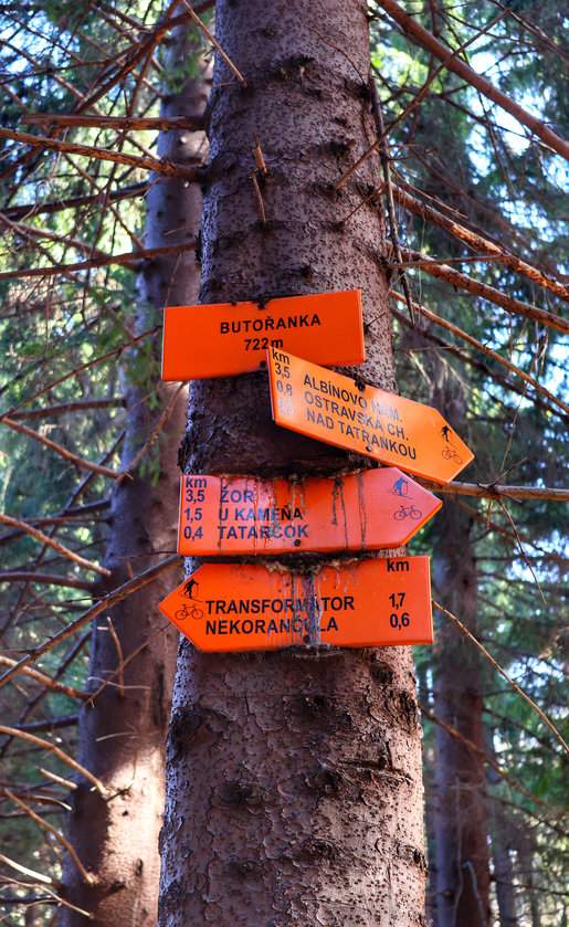 Pomarańczowa tablica z napisem ‘Butořanka 722 m’ zawieszona na drzewie, obok drogowskazy
