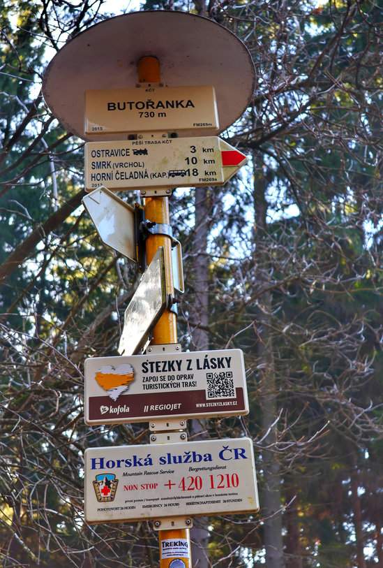 Biała tablica z napisem ‘Butořanka 730 m’ zawieszona na drzewie, obok drogowskazy